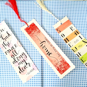 May include: Three handmade bookmarks with colorful watercolor designs and tassels. The first bookmark features a pink and white watercolor background with the text "you had the power all along my dear - Glinda". The second bookmark has a red and white watercolor background with red glitter and the text "home there's no place like...". The third bookmark has a rainbow watercolor background with the text "Somewhere over the rainbow".
