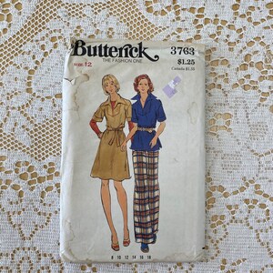 May include: A vintage sewing pattern for a dress and trousers. The pattern is from Butterick, the fashion one, and is size 12. The pattern number is 3763 and the price is £1.25. The pattern features two women wearing the garments. The dress is a brown, belted, short-sleeved dress. The trousers are plaid and have a blue waistband.