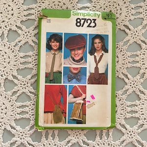 May include: Simplicity sewing pattern number 8723 for a one-size top, hat, and bag. The pattern cover features three models wearing different variations of the design. The models are wearing a brown, white, and red top with a brown tie, a brown sweater with a brown bow tie, and a white top with a brown tie.