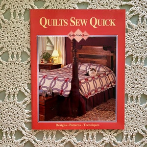 以下が含まれることがあります： 「キルト ソー クイック」というタイトルの赤い本で、副題は「キルトを簡単に」。表紙には、リング模様のキルトをかけたベッドのある寝室の写真が掲載されています。本には「デザイン - パターン - テクニック」とも書かれています。