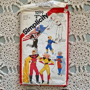 Puede incluir: Un paquete de patrones de costura Simplicity vintage con ilustraciones de disfraces de superhéroes. El paquete es blanco con detalles en rojo e incluye el texto "Simplicity" y "5299". Talla mediana, para adultos, talla 91-97 cm.