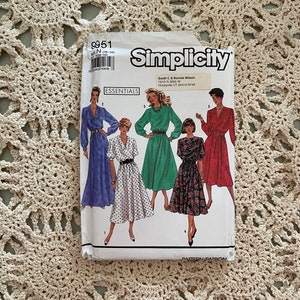 May include: Simplicity sewing pattern number 9951 for sizes 10-14. The pattern includes five different dress styles: a blue dress with long sleeves, a white dress with black polka dots and long sleeves, a green dress with long sleeves, a floral print dress with long sleeves, and a red dress with long sleeves. The pattern is labeled "Essentials".