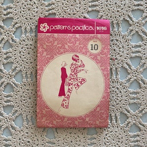 Puede incluir: Un patrón de costura Patterns Pacifica vintage, talla 10, con un diseño floral rosa y blanco. El paquete del patrón tiene un fondo rosa con el nombre de la marca y un dibujo de una mujer con un mono floral. El paquete está sobre un tapete de ganchillo color crema.