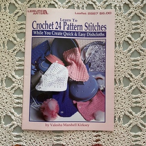 Puede incluir: Un folleto de crochet rosa y blanco titulado "Learn To Crochet 24 Pattern Stitches" con el subtítulo "While You Create Quick & Easy Dishcloths". El folleto presenta una foto de varios paños de cocina de crochet y una olla azul.