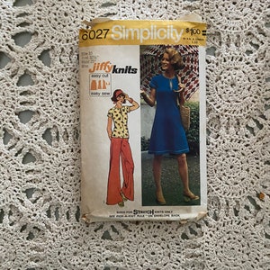 May include: Simplicity sewing pattern number 6027 for a dress and trousers. The pattern is for stretch knits only. The pattern cover features a woman wearing a blue dress and a woman wearing a red and white top and trousers.