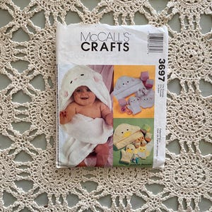 Op de afbeelding: Een McCall's Crafts patroon envelop met een baby in een witte badhanddoek met capuchon. Het patroon bevat afbeeldingen van babyaccessoires. De tekst "MCCALL'S CRAFTS" staat bovenaan. Het patroonnummer is 3697.