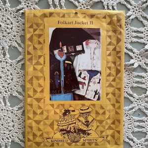 Könnte beinhalten: Ein Folkart Jacket II-Schnittmusterbuch mit einem goldenen geometrischen Rand. Das Cover zeigt ein Foto von mehreren verzierten Jacken und die Worte "Kindred Spirits". Der Hintergrund ist eine cremefarbene Häkelspitze.