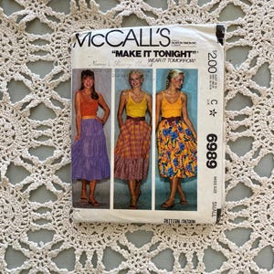 May include: A vintage McCall's sewing pattern packet featuring three women modeling tiered skirts and tank tops. The packet reads "MAKE IT TONIGHT" and includes the pattern number 6989. The background is a cream-colored, open-weave textile.