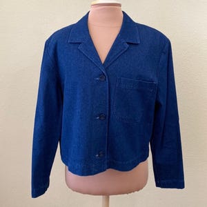 May include: A dark blue denim jacket with a collared neckline, three buttons, and a single pocket on the chest. The jacket is displayed on a mannequin and has long sleeves.