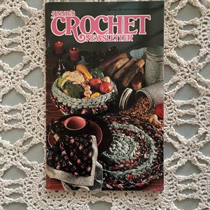 May include: A vintage copy of "Annie's Crochet Newsletter" magazine. The cover features the title in pink and green, with images of crocheted items and food. The magazine is set on a crocheted doily.