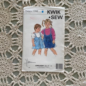 May include: A sewing pattern for a girls' dress with a blue and white gingham print. The pattern is from Kwik Sew Pattern Co., Inc. and is for sizes 4-5-6-7. The pattern shows two girls wearing the dress, one with a pink shirt and the other with a blue shirt.