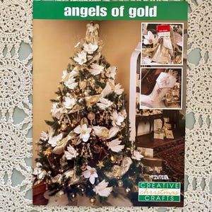 May include: A Christmas tree decorated with gold ribbon, white magnolia flowers, and gold baubles. The tree is topped with a gold angel figurine. The image includes the text "angels of gold" and "Leisure Arts Creative Christmas Crafts".