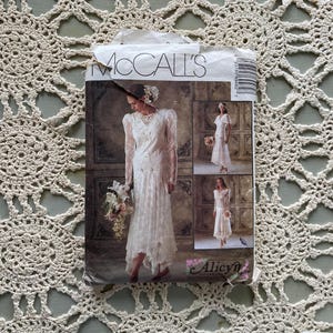 May include: A vintage McCall's sewing pattern for a wedding dress. The pattern shows a woman in a long, white lace gown with long sleeves and a high neckline. Additional images show variations of the dress. The pattern is labelled "Alicyn".