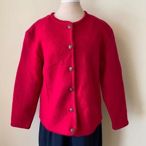May include: A bright red cardigan sweater with a round neckline and long sleeves. The sweater has a textured appearance and is fastened with five round, silver-colored buttons down the front. The hemline and neckline have a scalloped edge.