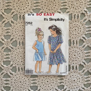 May include: A Simplicity sewing pattern for a girl's dress. The pattern number is 7252 and the size is 4-8. The pattern shows two girls wearing the dress, one in a solid blue and the other in a floral print.
