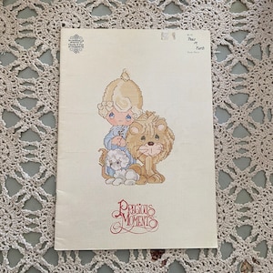 May include: A cross-stitch pattern featuring a Precious Moments character with a lion and a lamb. The pattern is titled "Peace on Earth" and is from Book PM 4.