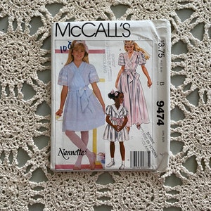 Puede incluir: Un patrón de costura McCall's vintage para vestidos de niña, con ilustraciones de las prendas terminadas. El paquete del patrón muestra la marca "Nannette" y el número 9474. La portada muestra un vestido azul claro y un vestido rosa.