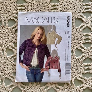Può includere: Un modello di cucito McCall's, M5243, con una donna che indossa una giacca viola fantasia. Il modello include opzioni di taglia EE (14-16-18-20). L'immagine è un primo piano del modello con il logo McCall's.