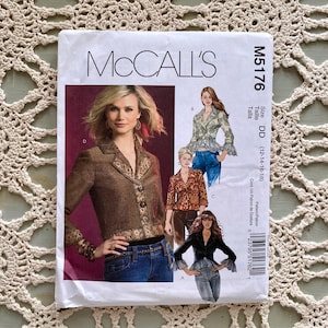 May include: A McCall's sewing pattern packet featuring a woman in a brown jacket with decorative trim. The packet includes images of different jacket styles and the text "M5176" and "Size DD (12-14-16-18)". The packet is on a cream-colored, crocheted surface.