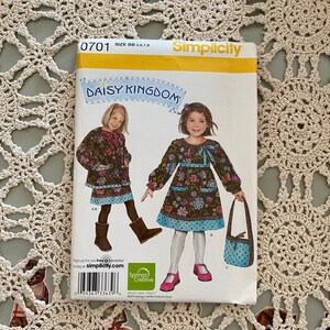 May include: A Simplicity sewing pattern for a girl's dress and matching bag. The pattern is for sizes BB 5, 6, 7, and 8. The dress is a brown floral print with a blue polka dot trim. The bag is a blue floral print with a brown trim. The pattern is titled "Daisy Kingdom".