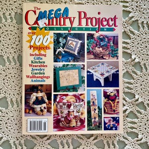 May include: The Mega Country Project Collection magazine cover features over 100 craft projects, including gifts, kitchen items, wearables, jewelry, garden decor, wall hangings, and animals. The cover shows a variety of crafts, including a pillow, a bird's nest, a picture frame, a stool, a doll, a planter, a wooden sign, and a basket.