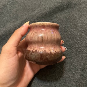 Puede incluir: Un pequeño jarrón de cerámica con forma ondulada y redondeada. El jarrón está esmaltado en un esquema de colores rosa y marrón con una base marrón más oscura.