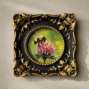 May include: A small square painting featuring a bumblebee on a pink clover flower, set within an ornate black frame with gold-painted decorative scrollwork accents along the edges and a circular inner border surrounding the artwork.