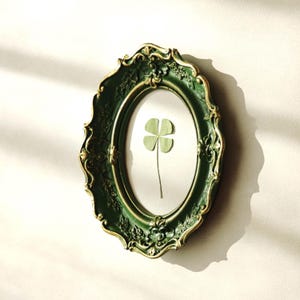 May include: An oval-shaped wall frame with a dark green finish and gold-toned highlights, featuring ornate floral relief detailing. Inside the frame, a single pressed four-leaf clover is centered against a plain, light-colored background.