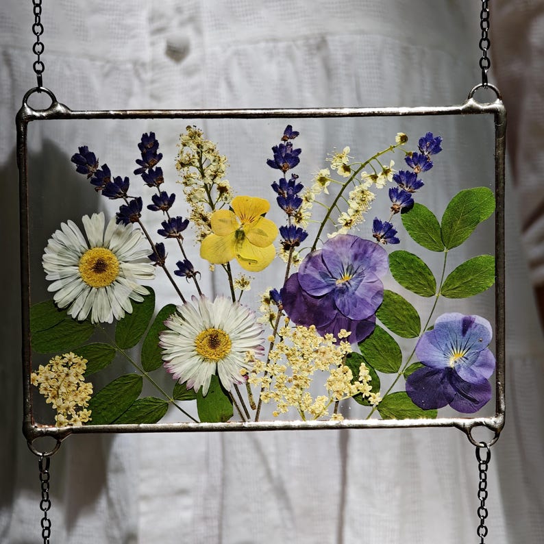 May include: A rectangular glass frame with a variety of dried flowers, including white daisies, yellow pansies, and purple lavender. The frame is edged with a silver metal and hangs from a chain.