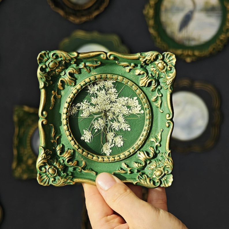 May include: A square, ornate green and gold frame featuring a circular center display containing a pressed white Queen Anne's lace flower. The frame has intricate, raised scrollwork details along its edges and a deep green matte finish.