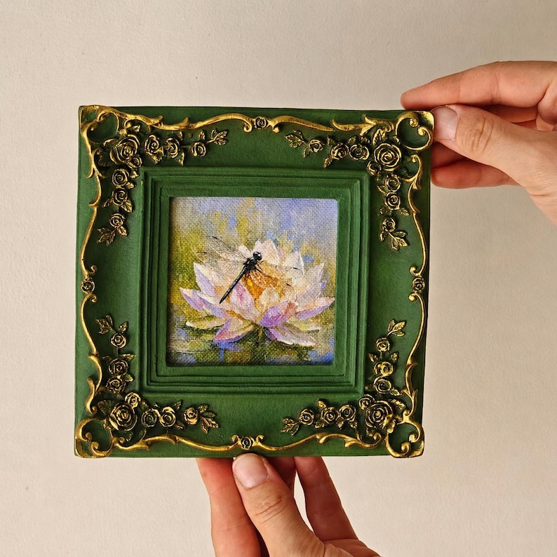 May include: A square painting of a dragonfly resting on a white and purple water lily, encased in a dark green frame with ornate gold-colored rose relief detailing on the corners and edges, featuring a textured finish on the frame surface.