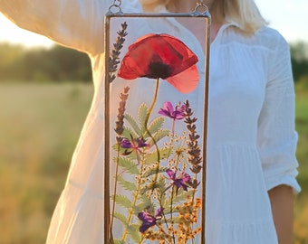 May include: A pressed flower art piece in a glass frame. The piece features a red poppy flower with purple and white wildflowers. The frame is hung on a white wall.