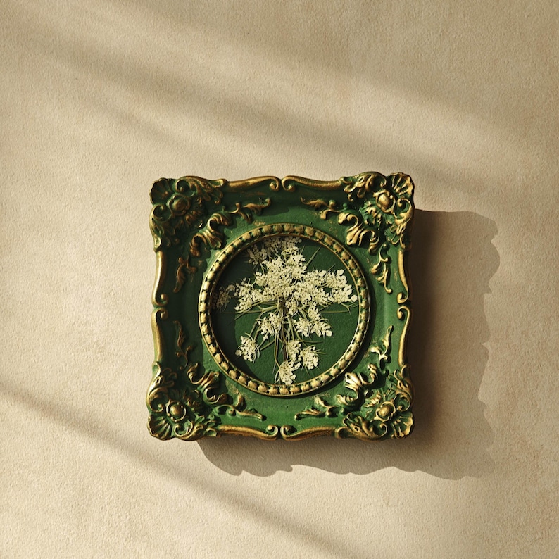 May include: A square wall frame with an ornate, dark green and gold finish featuring a circular center cutout. Inside the circular opening, a pressed white wildflower arrangement is displayed against a matching dark green background.