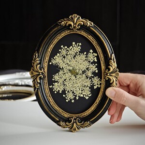 May include: An oval black and gold framed pressed flower art piece. The frame has ornate detailing and a black background. The pressed flower is a light cream colour and is centered in the frame. The frame is being held by a hand.