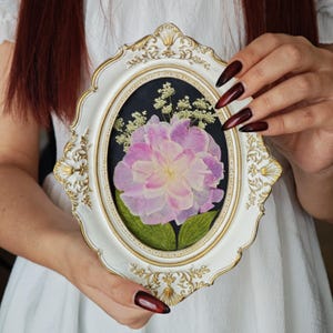 May include: An oval, white and gold framed pressed flower art piece. The artwork features a large, light pink and white flower with green leaves against a dark background. The frame has ornate gold detailing.