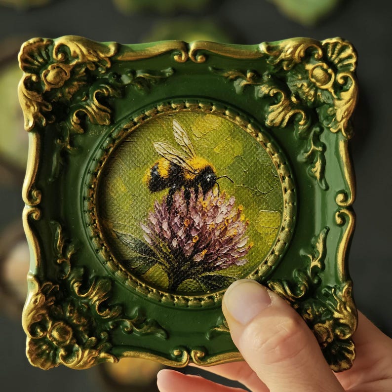 May include: A small square frame with an ornate dark green and gold border surrounding a circular painting. The artwork depicts a fuzzy bumblebee perched on a textured purple flower against a green background, rendered with visible brushstrokes.