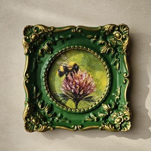 May include: A square wall art piece featuring a circular painting of a bumblebee on a pink flower. The frame is dark green with ornate gold-painted relief details at the corners and edges, surrounding the textured canvas artwork.