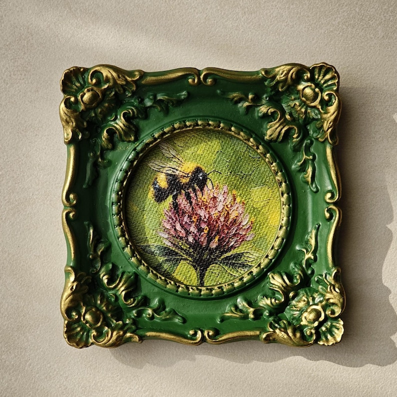 May include: A square wall art piece featuring a circular painting of a bumblebee on a pink flower. The frame is dark green with ornate gold-painted relief details at the corners and edges, surrounding the textured canvas artwork.