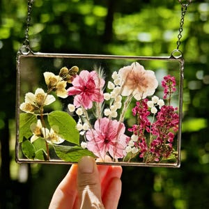 May include: A rectangular glass art piece with pressed flowers. The arrangement includes pink, white, and yellow flowers, with green leaves. The piece is suspended by a silver chain. The background is blurred green.