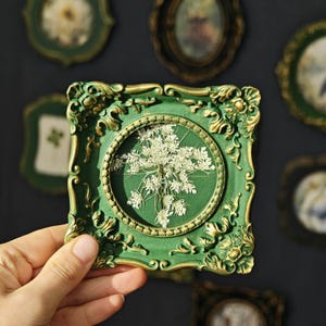 May include: A square picture frame with an ornate, embossed green and gold border. The frame features a circular center containing a pressed white Queen Anne's lace flower against a solid green background. The design includes intricate scrollwork.