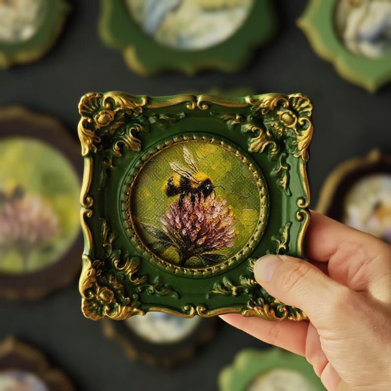 May include: A small square frame with ornate gold-painted scrollwork details on a dark green base. The frame holds a circular painting of a bumblebee resting on a textured pink clover flower, rendered with visible brushstrokes and earthy tones.