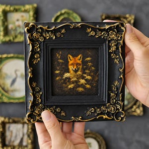 May include: A square picture frame in matte black with raised gold-painted rose and vine detailing. The frame contains a dark-toned print of a fox surrounded by golden-hued foliage and flowers, set against a textured, dark background.