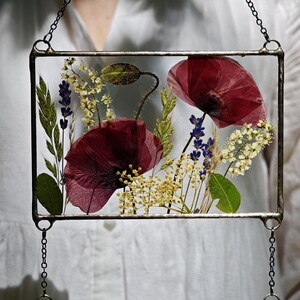 May include: A rectangular glass frame with pressed flowers, including deep red poppies, lavender, and yellow blooms. The frame is suspended by a chain. The flowers are arranged against a clear background, creating a delicate, botanical display.