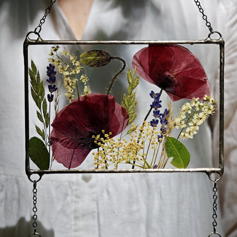 May include: A rectangular glass frame with pressed flowers, including deep red poppies, lavender, and yellow blooms. The frame is suspended by a chain. The flowers are arranged against a clear background, creating a delicate, botanical display.