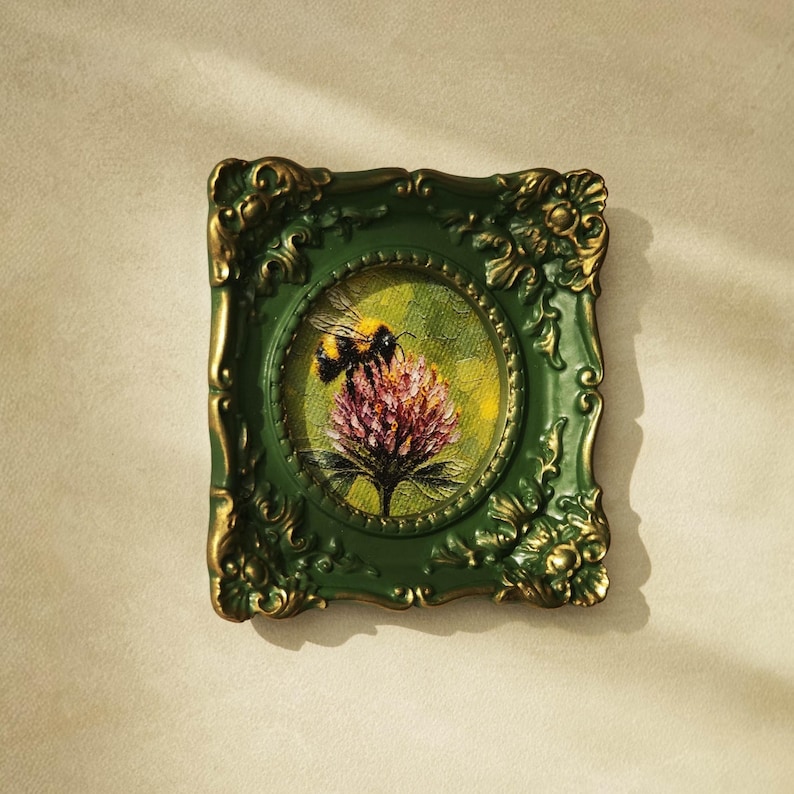 May include: A small rectangular wall frame with an ornate, dark green and gold finish. The frame surrounds a circular painting of a bumblebee perched on a pink clover flower against a textured yellow and green background with visible brushstrokes.