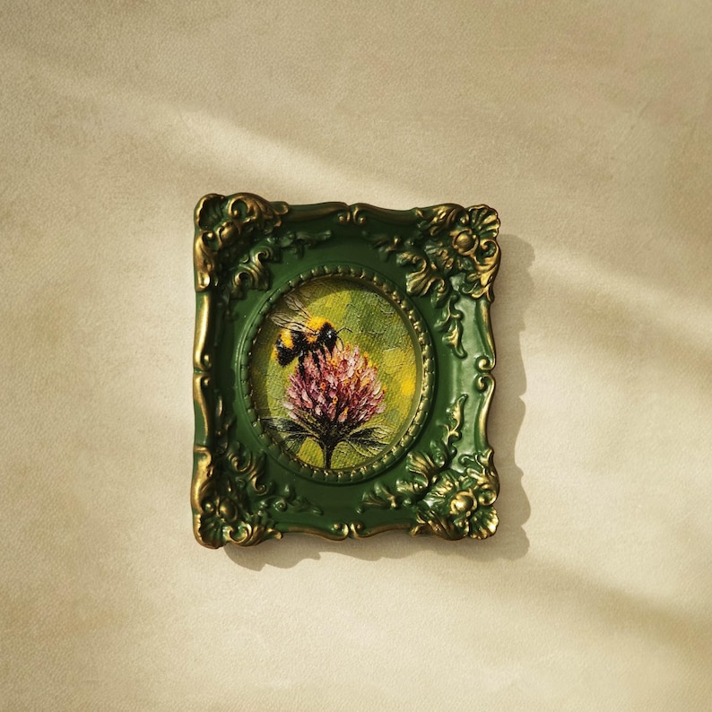 May include: A small rectangular wall frame with an ornate dark green and gold finish, featuring a circular inner border. Inside the frame is a painting of a bumblebee resting on a pink clover flower against a textured yellow and green background.