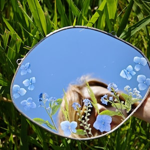 Może przedstawiać: Okrągłe lustro ze srebrną ramą, ozdobione niebieskimi kwiatami i zielenią. Lustro odbija czyste, błękitne niebo. Lustro jest umieszczone na tle zielonej trawy.