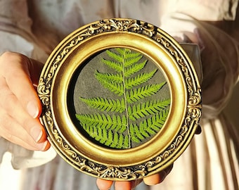 May include: A round, gold-colored frame with ornate detailing holds a vibrant green fern leaf against a dark background. The frame is held in front of a soft, neutral-toned background, highlighting the natural beauty of the botanical art piece.
