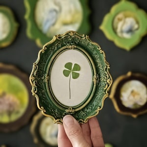 May include: An oval-shaped picture frame with a dark green finish and gold-toned decorative scrollwork edges. The frame contains a light-colored backing material featuring a single pressed four-leaf clover centered within the oval display area.
