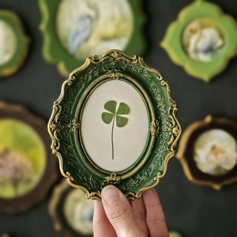 May include: An oval-shaped picture frame with a dark green finish and gold-toned decorative scrollwork edges. The frame contains a light-colored backing material featuring a single pressed four-leaf clover centered within the oval display area.